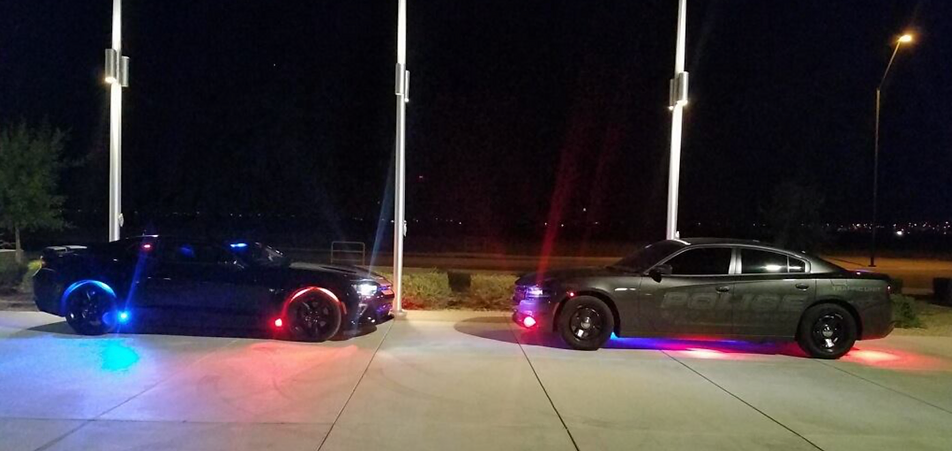 Two black police cruisers parked at night with headlights and underbody lights on